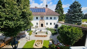 Ein weißes Gebäude mit einer Solaranlage auf dem Dach, einem zentralen Eingang mit dekorativem Bogen und einem Steinfbrunnen im Innenhof.