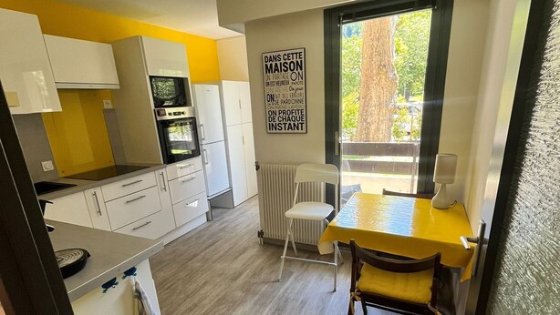 Cocina compacta con armarios blancos, una mesa de comedor amarilla y una ventana con vistas a la vegetación.