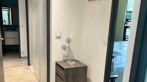 Un pasillo con piso de madera, un mueble pequeño de madera con cajones, y una lámpara fijada en la pared.
