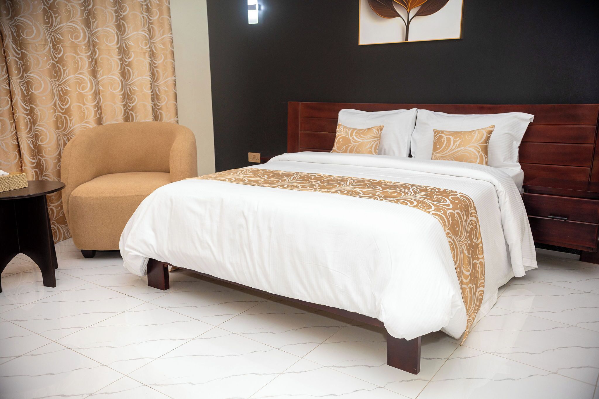 A neatly made bed with a white and gold bedspread, a brown upholstered armchair, and a dark wooden nightstand with a lamp.