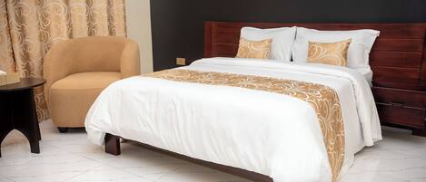 A neatly made bed with a white and gold bedspread, a brown upholstered armchair, and a dark wooden nightstand with a lamp.