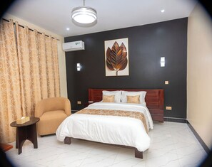 A hotel room with a bed, a chair, a small table, an air conditioning unit, and a wall-mounted artwork. - DEEL Comfort Inn (Dar es Salaam)