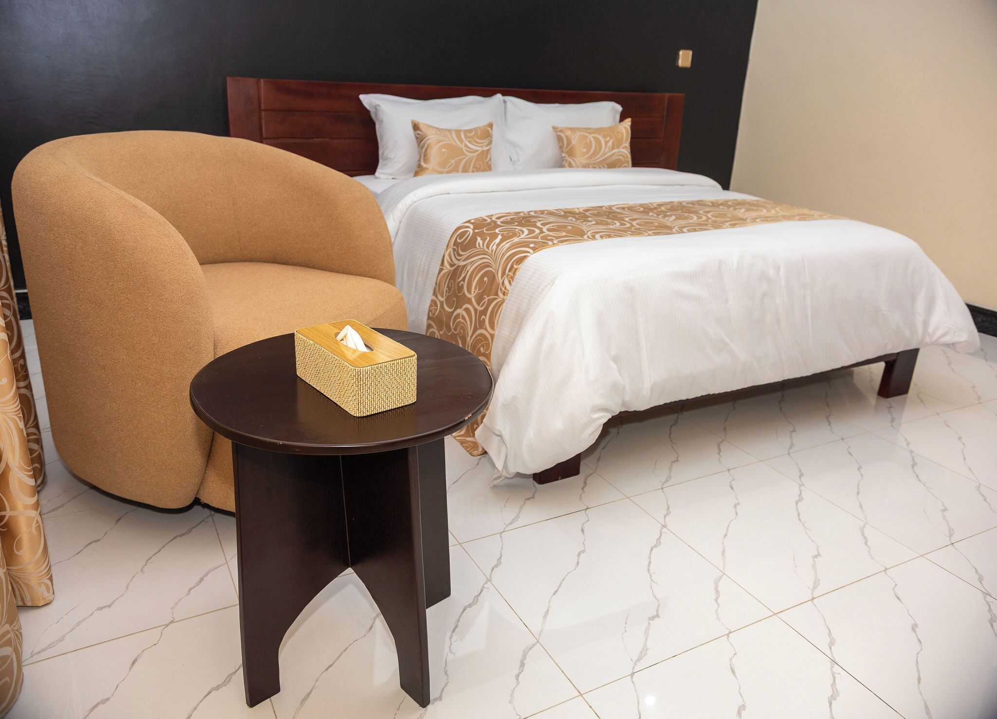 A hotel room with a large armchair, a bedside table with a tissue box, a neatly made bed with patterned bedding, and a marble-tiled floor.