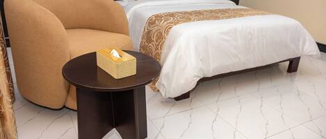 A hotel room with a large armchair, a bedside table with a tissue box, a neatly made bed with patterned bedding, and a marble-tiled floor.