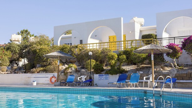 Una piscina con acqua blu, circondata da edifici bianchi con porte gialle e lettini blu.