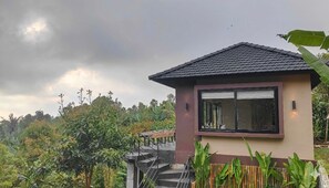 A small, modern house with a tiled roof and a large window. It is surrounded by lush greenery and has a staircase leading up to it.