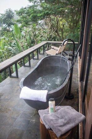 A stone bathtub filled with water, a towel, and a bottle on a wooden stool, with a wooden railing and greenery in the background.