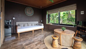 Ein modernes Wohnzimmer mit einer Couch, einem runden Tisch mit Hocker, einem Deckenventilator und Blick ins Grüne durch ein Fenster.