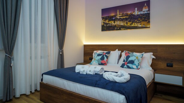 Una cama bien tendida con colcha azul, almohadas blancas y un cuadro de paisaje urbano sobre ella.