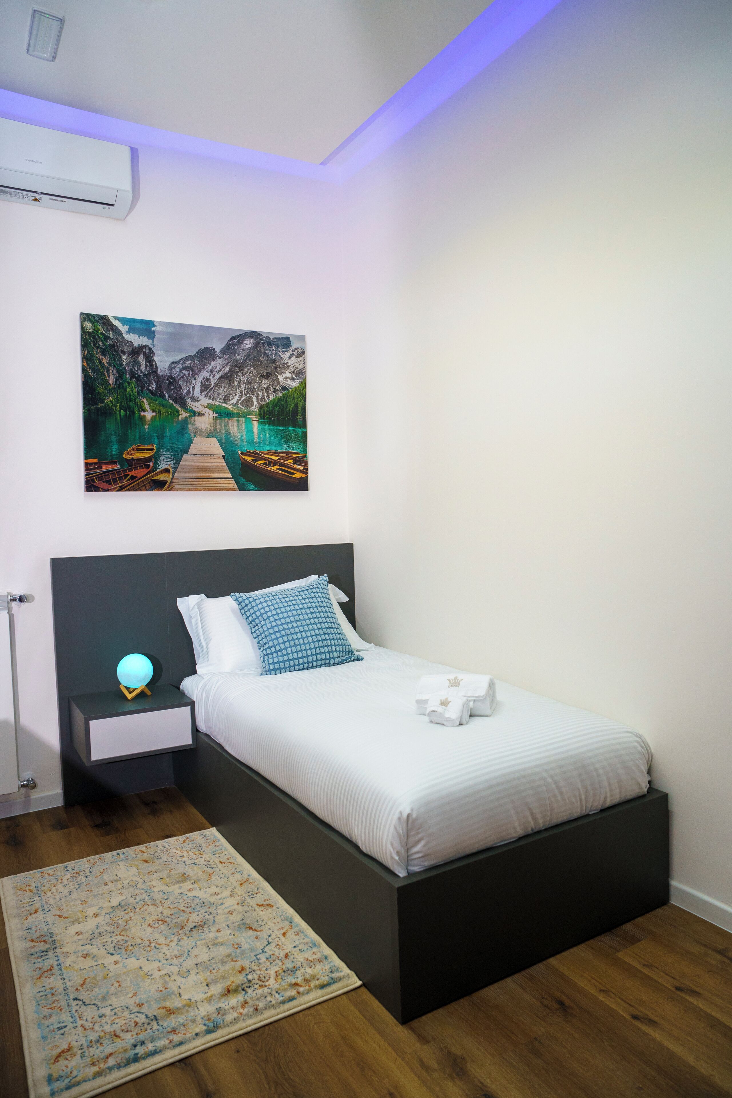 A bedroom with a bed, a nightstand, a wall-mounted air conditioner, a painting, and a floor lamp.