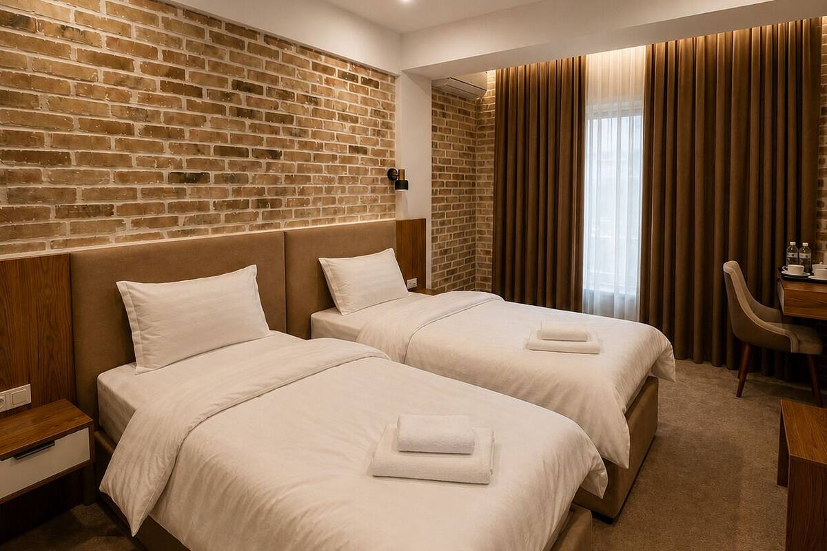 A hotel room with two beds, a brick wall, a wooden desk, and a window with curtains.