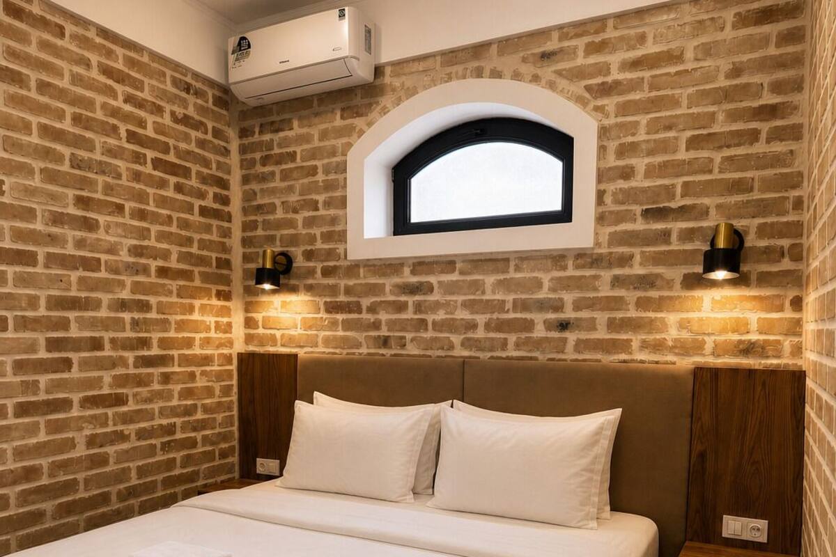 A hotel room with a brick wall, a bed with white linens, a small window, and two bedside lamps.
