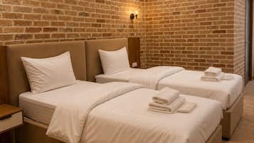 Habitación de hotel con pared de ladrillo, dos camas con sábanas blancas, una mesita de noche y una lámpara fijada a la pared.