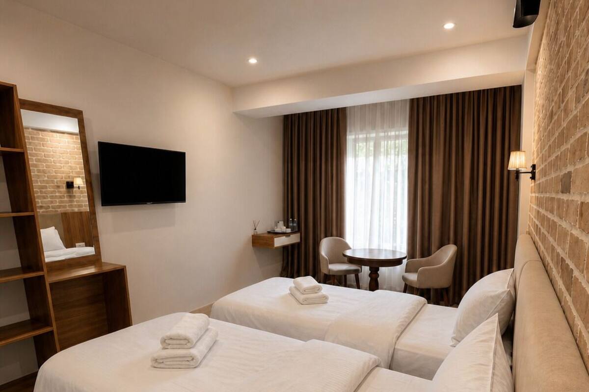 A hotel room with two beds, a TV, a desk with a chair, and a brick wall.