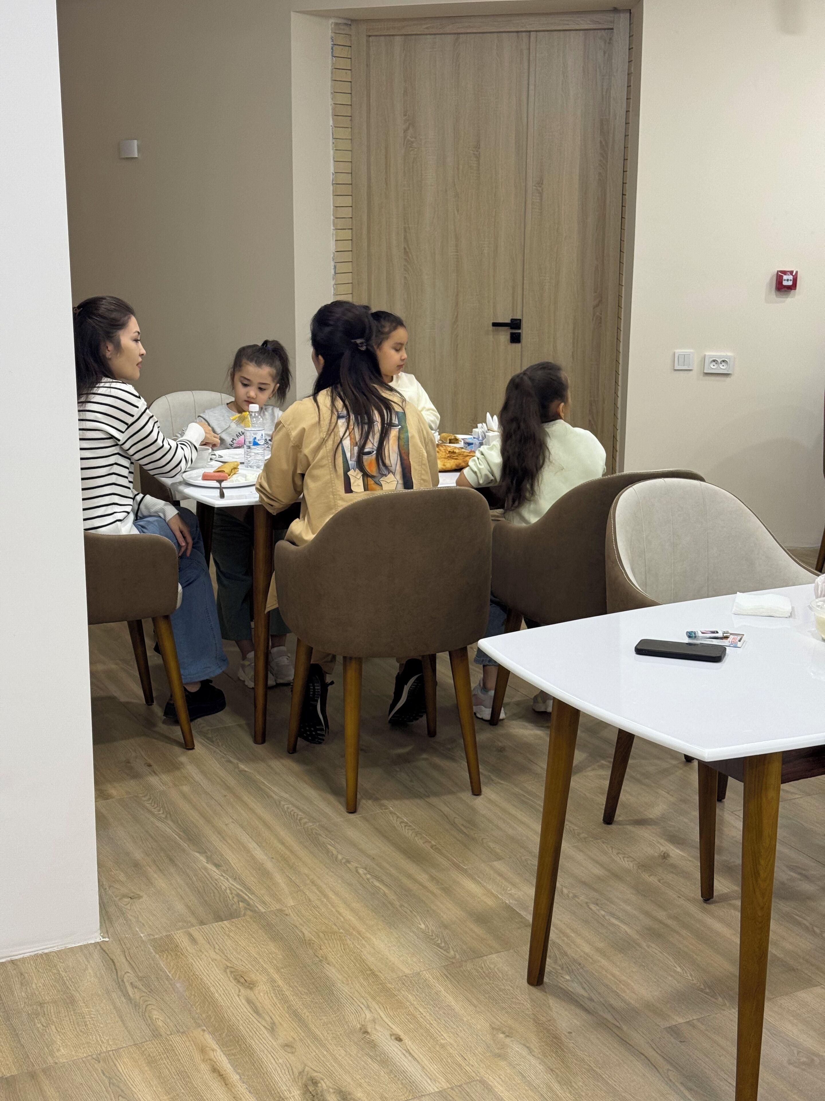 A group of people seated at a round table in a room with a wooden door and light-colored walls.