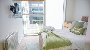 Une chambre avec une grande fenêtre, un lit recouvert d’une couverture verte, un radiateur blanc et un téléviseur fixé au mur.