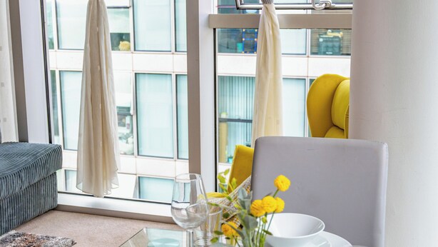 Un coin repas avec une table dressée pour deux personnes, un vase rempli de fleurs jaunes et une vue sur la ville, visible à travers de grandes fenêtres ornées de rideaux légers.