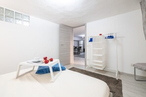 A modern room with a bed, a small table with a cup and flowers, a white wardrobe, and a door leading to a dining area.