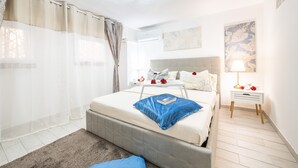 Une chambre à coucher comprenant un lit, une table de chevet, une commode, une lampe et une fenêtre avec des rideaux.