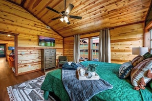 L’intérieur d’un chalet en bois comprend un lit, un ventilateur de plafond, un téléviseur et une fenêtre avec des rideaux.