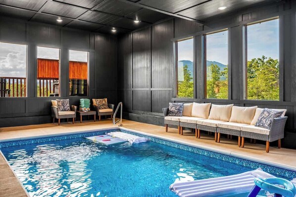 Une piscine intérieure avec un canapé et des fauteuils, de grandes fenêtres et une vue sur la verdure.
