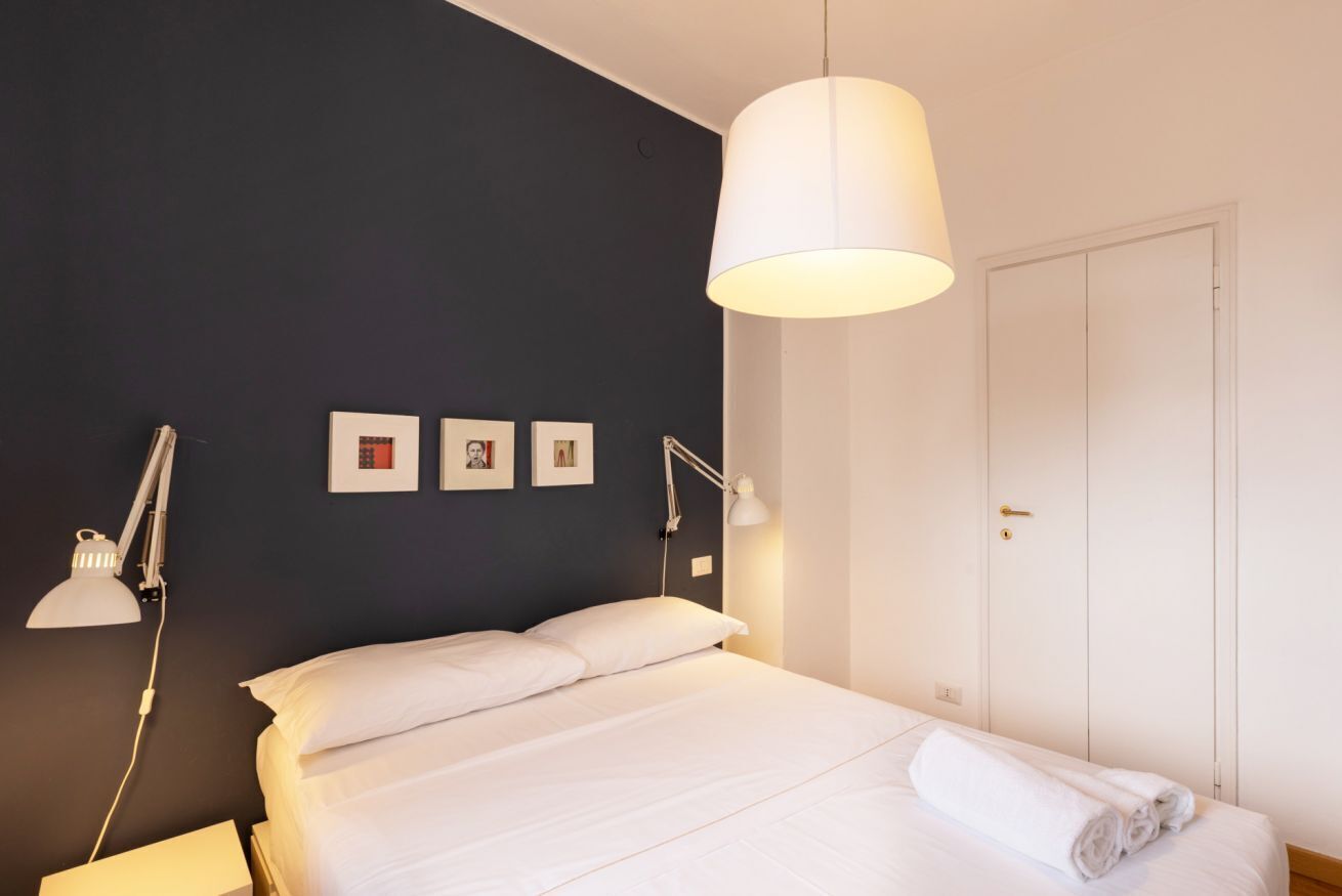 Una cama bien hecha con ropa blanca, una lámpara de noche y una lámpara de pared. Hay tres cuadros enmarcados en la pared.