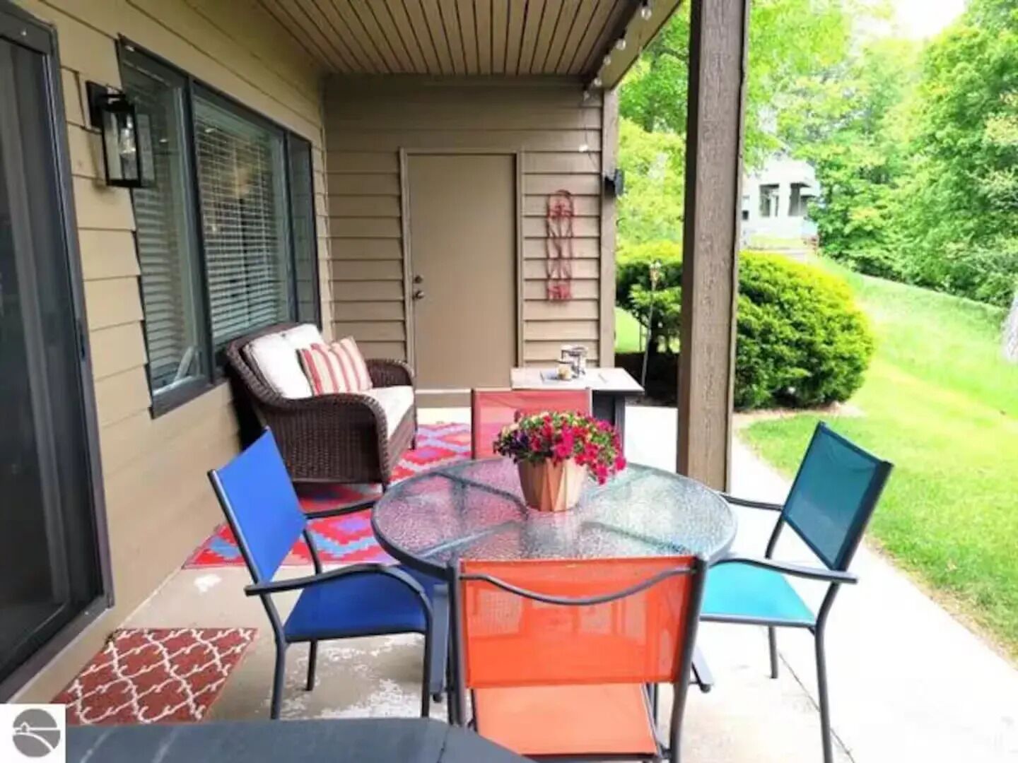 A covered patio with a glass table, blue and orange chairs, a wicker sofa, and a potted plant.