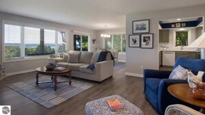A spacious living room with a sofa, coffee table, and ottoman. There is a kitchen area with a countertop and cabinets. - 2BR Escape Torch Lake Views Golf 755 (Kewadin)