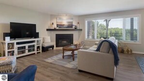 A living room with a flat-screen TV, a fireplace, a sofa, and a window with blinds. - 2BR Escape Torch Lake Views Golf 755 (Kewadin)