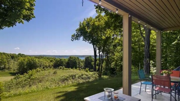 An outdoor patio with a view of a grassy field and trees, a table with a candle and a jar, and a covered seating area with chairs. - 2BR Escape Torch Lake Views Golf 755 (Kewadin)