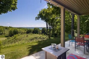 An outdoor patio with a view of a grassy field and trees, a table with a candle and a jar, and a covered seating area with chairs. - 2BR Escape Torch Lake Views Golf 755 (Kewadin)