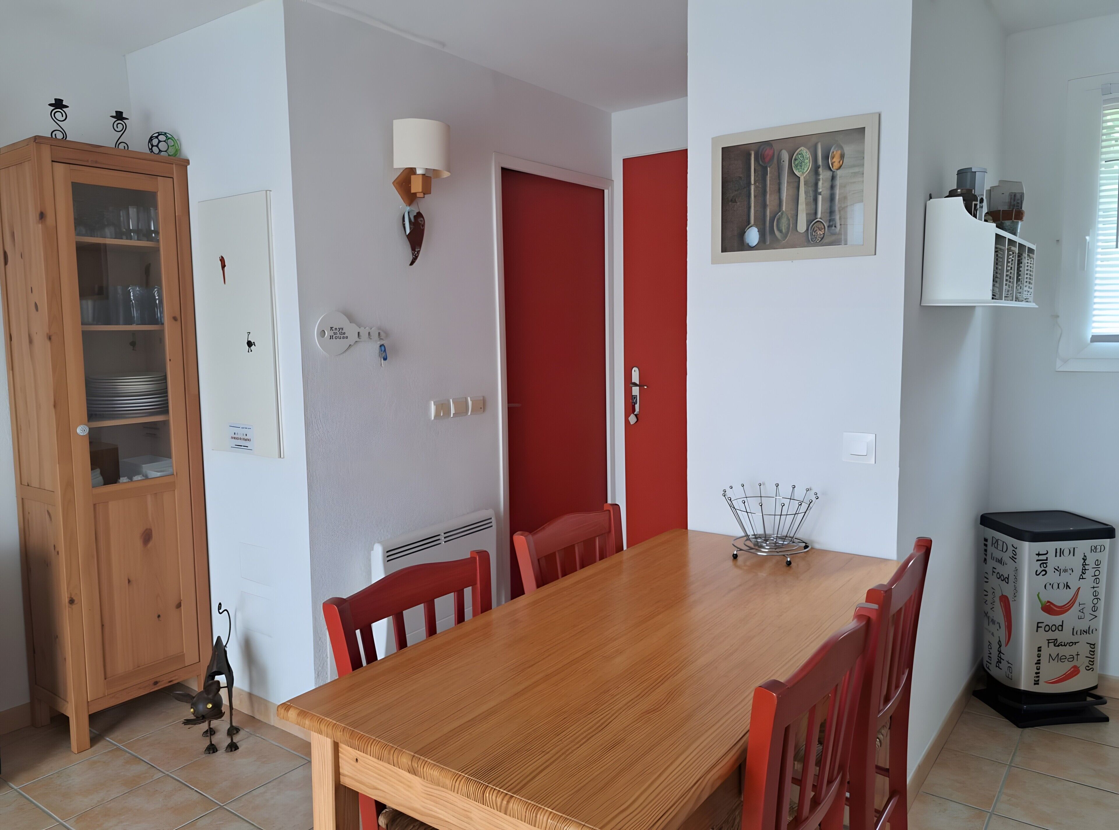 Een eetgedeelte met een houten tafel, rode stoelen, een kast met glazen deuren, een wandlamp en een lijst met afbeelding van keukengerei.