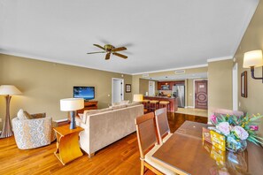 A living room with a sofa, a dining table, and a TV. There is a kitchen in the background.