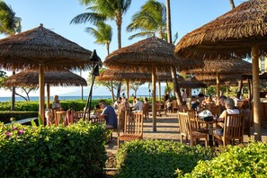An outdoor dining area with thatched umbrellas and wooden chairs, overlooking a beach.
