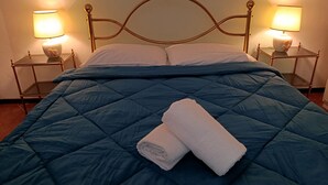 Un lit bien fait, recouvert d’une couette bleue, avec deux oreillers blancs et une serviette blanche pliée. Deux tables de chevet, chacune surmontée d’une lampe, se trouvent de part et d’autre du lit.