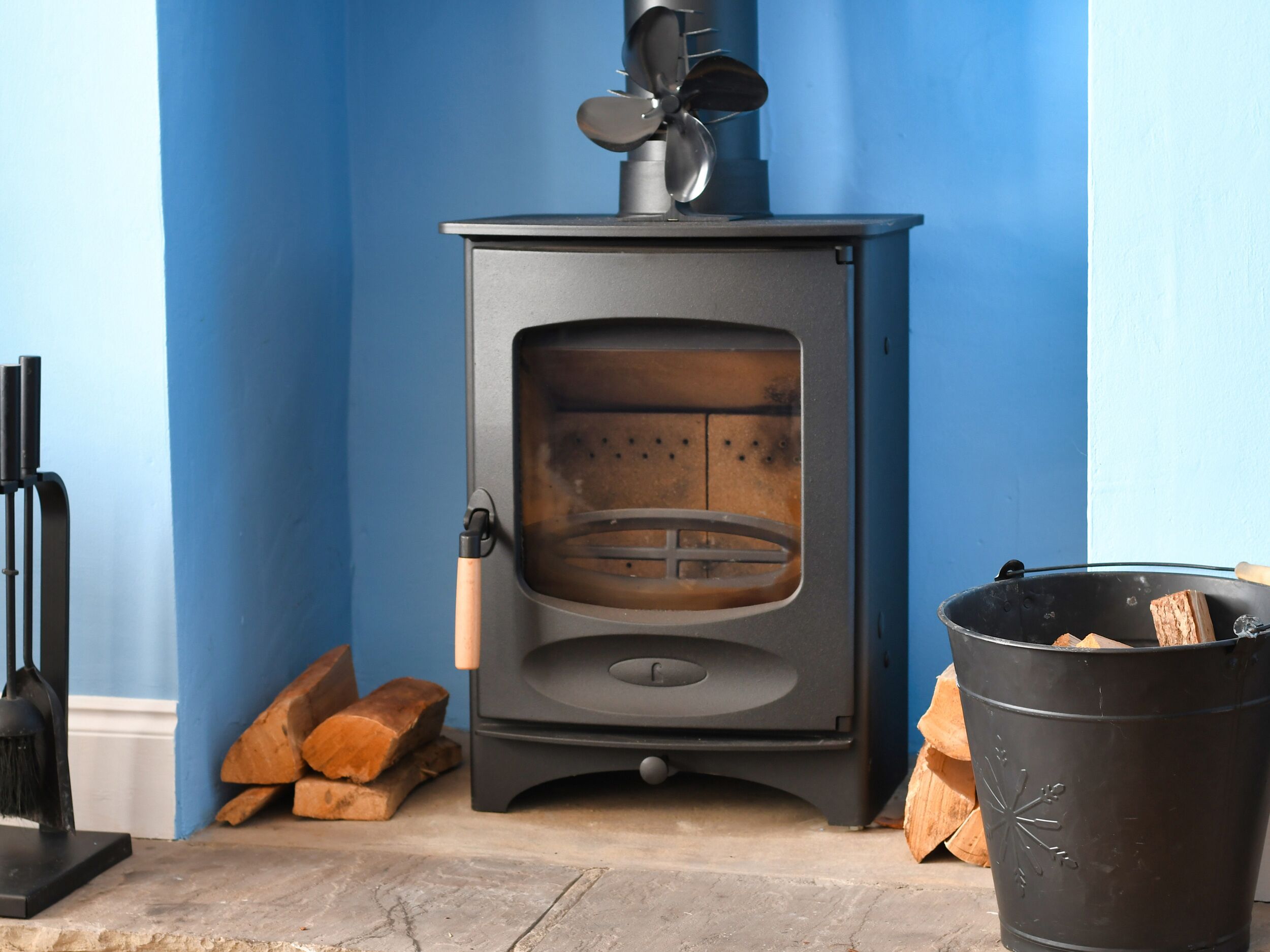 A wood-burning stove with a glass door, a chimney, and a bucket of firewood.