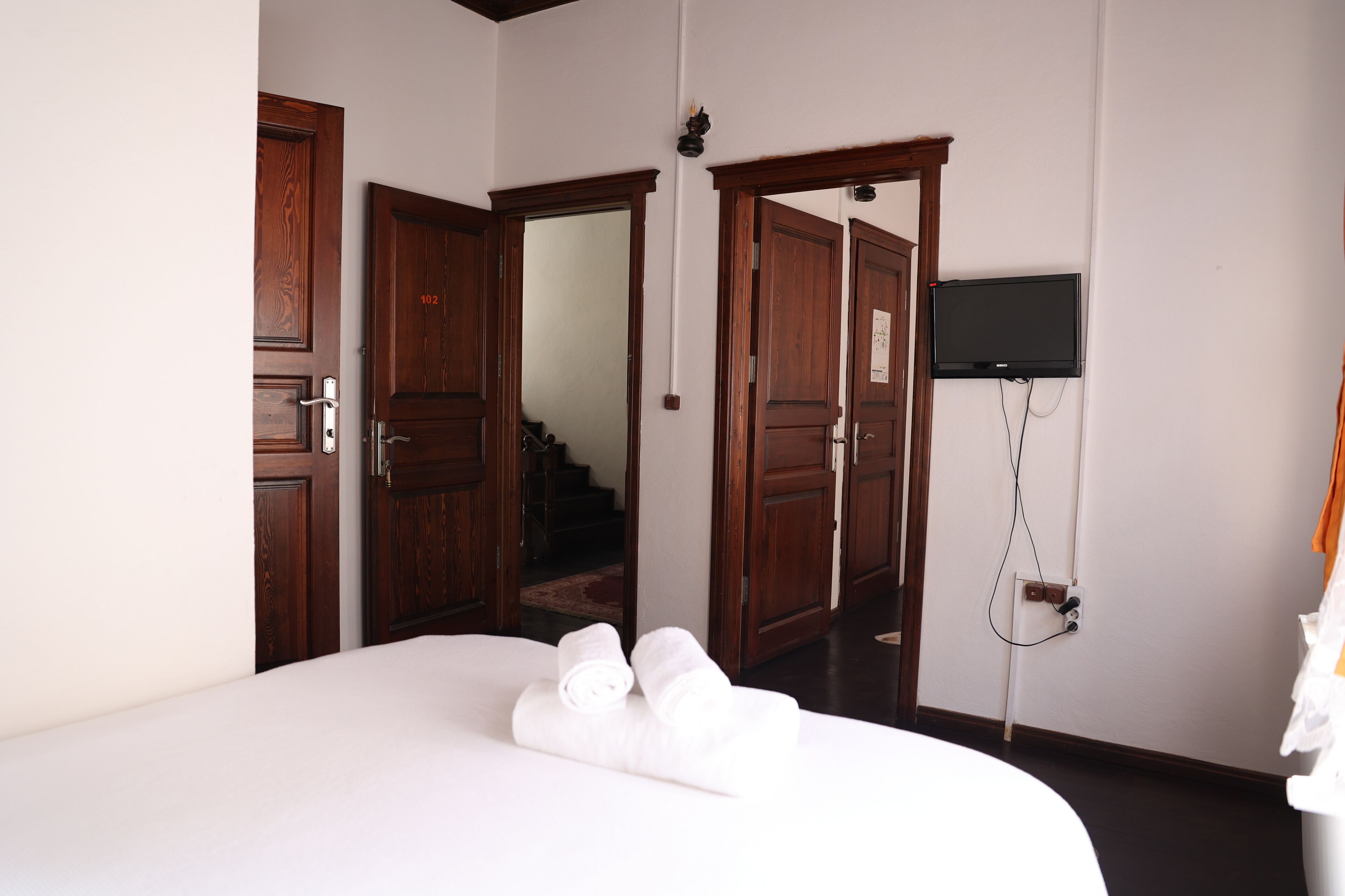 A hotel room with a large bed, wooden doors, a flat-screen TV mounted on the wall, and a visible electrical outlet.