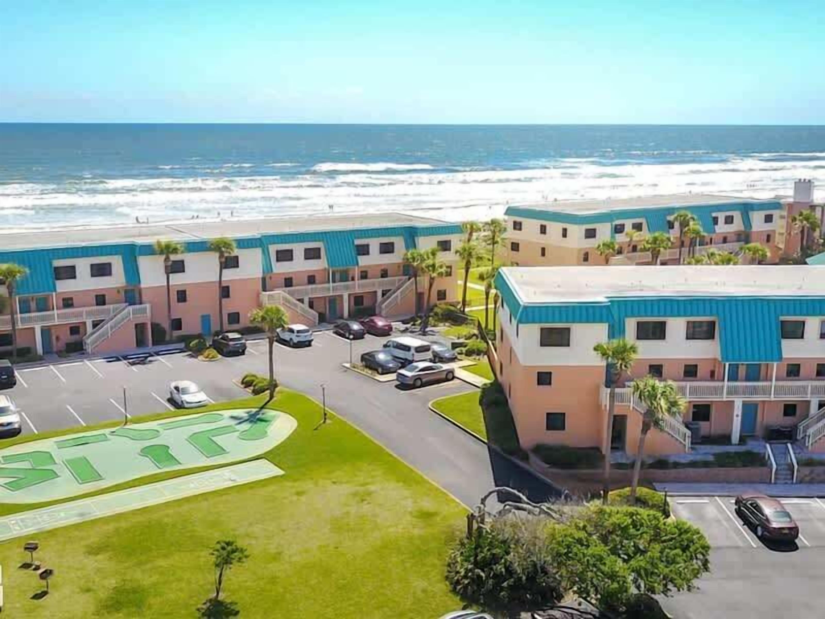 Un hotel frente a la playa con edificios de varios pisos, estacionamiento y vistas al océano.