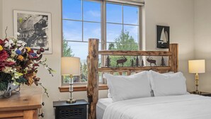 Ein Schlafzimmer mit Holz-Kopfteil, weißer Bettwäsche, einem Nachttisch mit Lampe, einem Schminktisch und einem Fenster mit Blick auf Bäume und ein Segelboot.