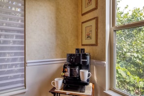 Una cafetera sobre una mesita con dos tazas, una ventana con persianas y un cuadro en la pared.
