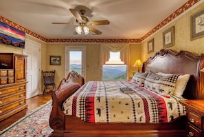 A bedroom with a large bed, a wooden dresser, a chair, a ceiling fan, and a view of the outdoors through a window.
