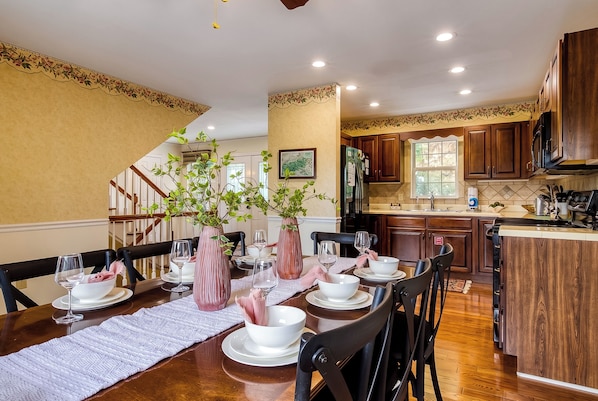 Un comedor con una mesa de madera dispuesta para una comida, rodeada de sillas, y una cocina al fondo.