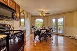 A kitchen with a stove, oven, and microwave. There is a dining table with chairs and a chandelier. The room has wooden floors and a view of trees through the windows.