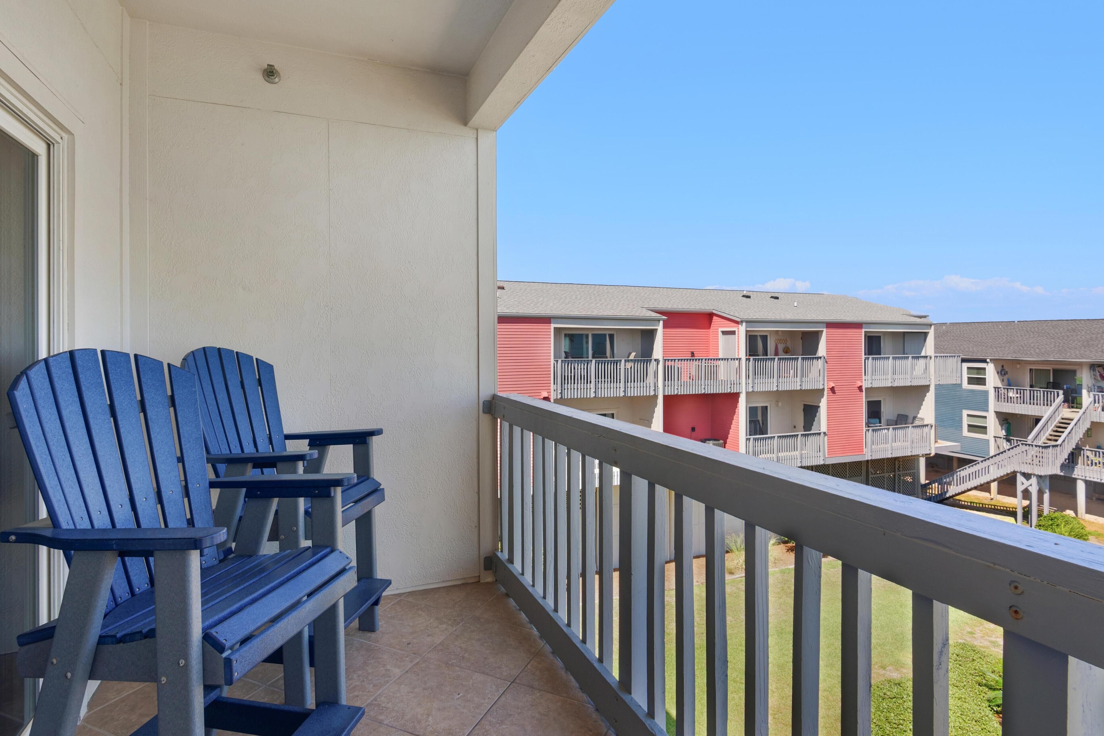 A balcony with two Adirondack chairs, a white railing, and a view of multi-story buildings with balconies and stairs.