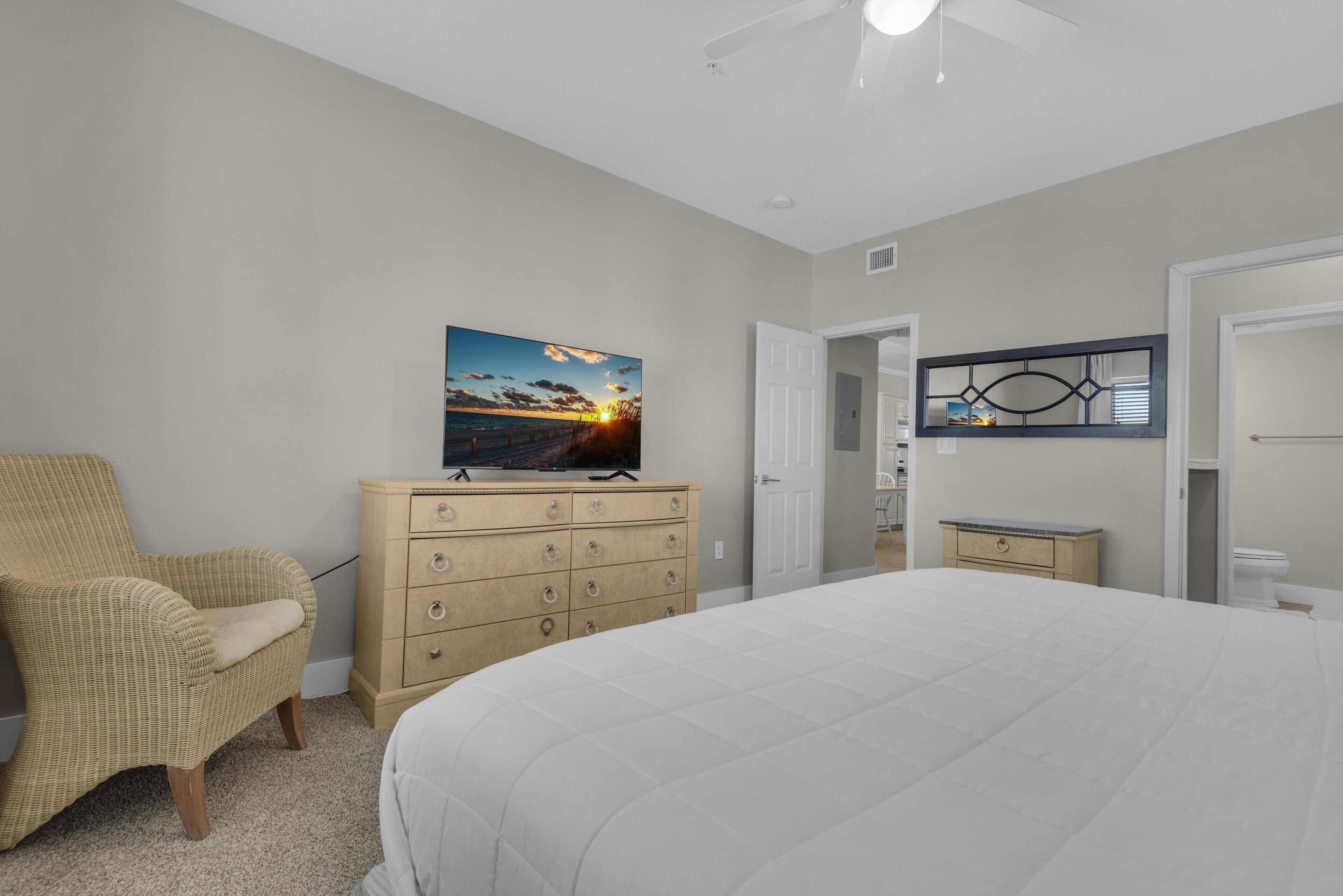 A bedroom with a large bed, a television on a dresser, a chair, and a door leading to another room.
