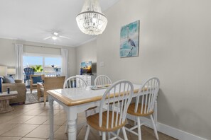 A dining area with a white table and chairs, a chandelier, and a painting of a bird on the wall. - Villas on the Gulf Unit J8 (Pensacola Beach)
