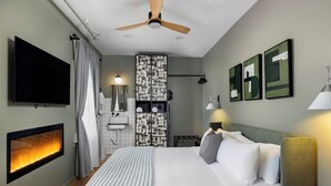 A hotel room with a bed, a flat-screen TV, a fireplace, a ceiling fan, and artwork on the walls. - Modal House: A Fun-Filled Designer Home Just Steps from Downtown Main Street (Greenville)