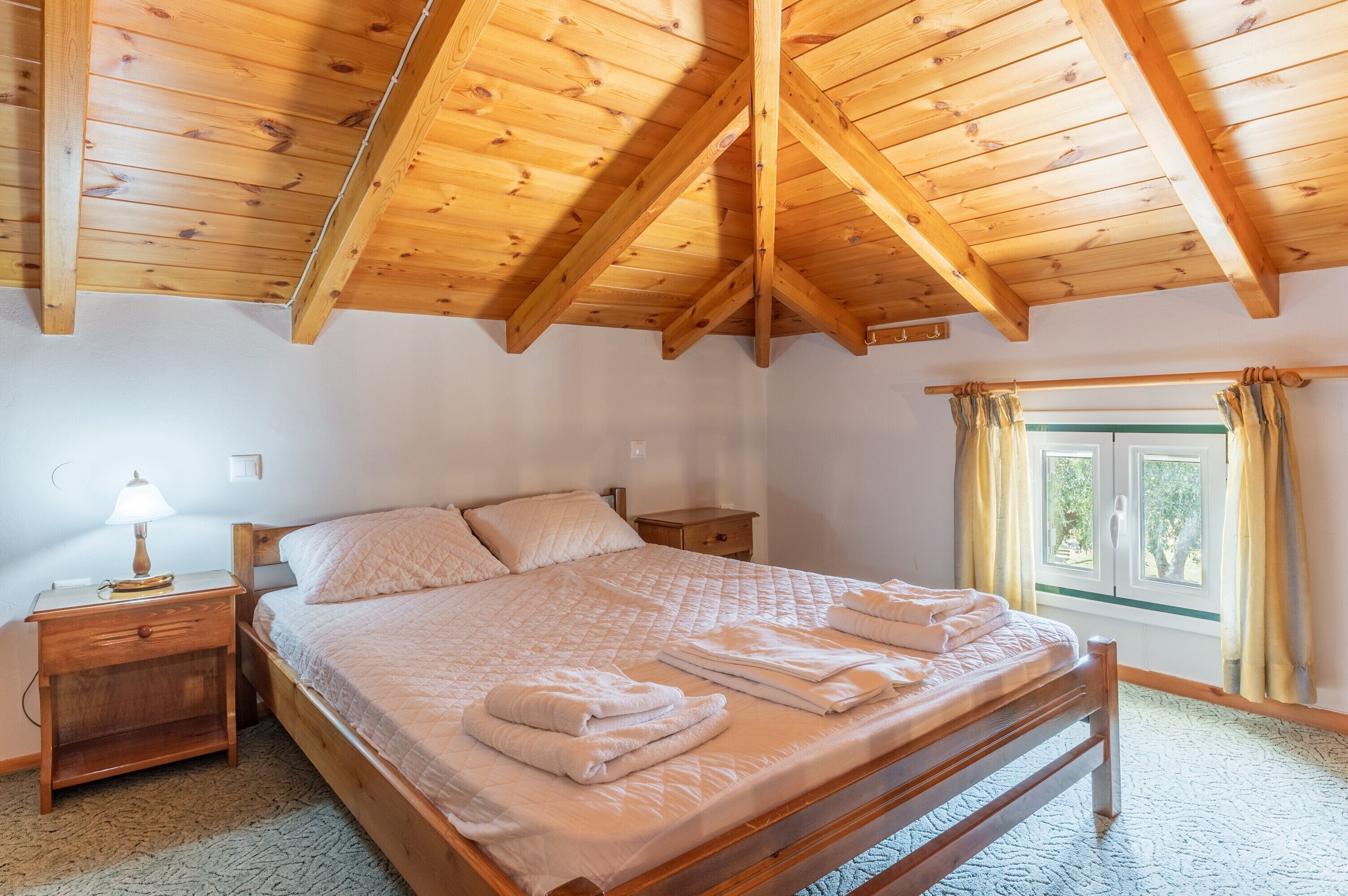 A bedroom with a wooden ceiling, a bed with pillows and a blanket, a nightstand with a lamp, and a window with curtains.