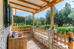 A covered outdoor seating area with stone walls and wooden benches, surrounded by a wooden fence and greenery.