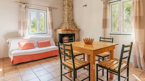 Una habitación acogedora con chimenea de piedra, una mesa de madera con sillas, un sofá con cojines y ventanas con cortinas.